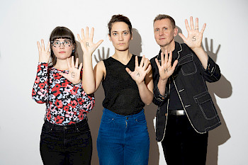 Zwei junge Frauen und ein Mann stehen vor einer weißen Wand und halten ihre Handinnenseiten in die Kamera, auf denen die Buchstaben G,R,A,M,S gesschrieben sind-