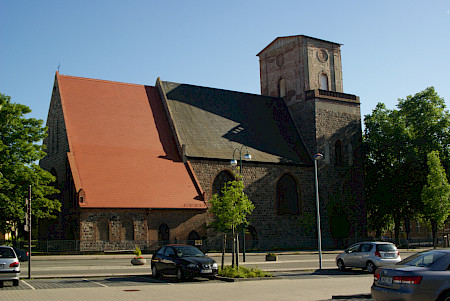 graue Kirche von der Seite, mit einem roten Dach, davor ein Parkplatz drei Autos