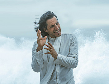 Ein Mann singt vor dem Meer, er hat blauen Anzug