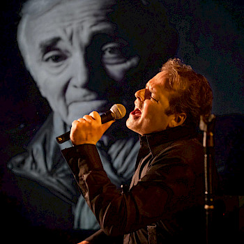 Mann singt leidenschaftlich in ein Mikrofon, hinter ihm eine Schwarz-Weiß-Projektion eines alten Mannes