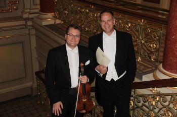 2 Männer im Frack stehen auf einer barocken Treppe, einer mit Noten, der zweite mit einer Violine in der Hand