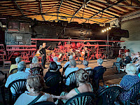 Blick über Publikum auf Plastikstühlen, die in einer Lokhalle auf Musiker vor einer historischen Dampflok schauen