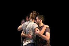 Zu sehen ist ein szenisches Bild aus einer Vorführung von "One More Thing". Vier Männer umarmen sich. Der Hintergrund ist schwarz.
