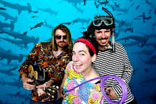 Die Band "Bungalow Gang" ist zu sehen. Sie tragen teilweise skurrile Accessoires. Im Hintergrund sieht man einen Schwarm Hammerhaie schwimmen.