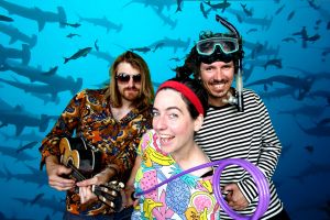 Die Band "Bungalow Gang" ist zu sehen. Sie tragen teilweise skurrile Accessoires. Im Hintergrund sieht man einen Schwarm Hammerhaie schwimmen.