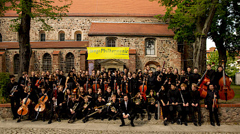 Zusehen ist das Junge Philharmonieorchester vor einer Kirche. Sie sind alle schwarzgekleidet und halten ihr Instrument in der Hand.