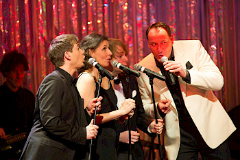 Drei Männer und eine Frau singen auf einer Bühne in Mikrofone, hinter ihnen ein festlich glitzernder Vorhang
