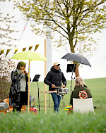 Zwei Musiker spielen unter schirmen draußen auf einer Wiese Flöte und Trommel. Im Vordergrund sitzt ein Zuschauer mit dem Rücken zur Kamera.