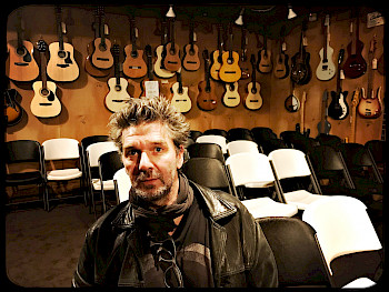 Der Komponist Helmut Oehring steht vor Stuhlreihen, an der Wand hängen Gitarren.