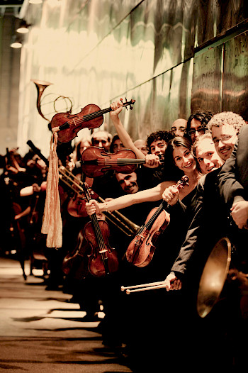 Ein Orchester: Die MusikerInnen sitzen nur an rechter Reihe. Aber sie jubeln und lachen zusammen.