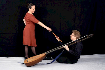 Theresia Stahl steht links in einem braun-rötlichen Kleid. Christian Stahl sitz rechts unten auf dem Boden mit seiner laute zu Frau Stahl gewandt und reicht ihr eine Blockflöte.