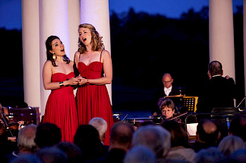 Zwei Opernsängerinnen stehen auf der Bühne nebeneinander. Sie tragen rote Kleider.