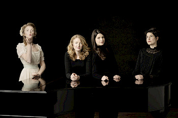 Vier Frauen stehen hinter einem Hammerklavier. Alle sind schwarz gekleidet, außer die Frau links außen. Sie trägt ein weißes Kleid.