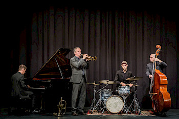 Luna Jazz Band spielt auf einer Bühne. Von links nach rechts Klavierspieler, Trompetenspieler, Schlagzeuger und rechts außen der Kontrabassist.