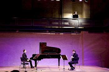 Zeigt ein Klavier Flügel und zwei Männer vor einem lila Hintergrund