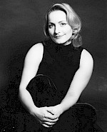 Ein schwarz-weißes Foto von Doerthe Maria Sandmann. Sie trägt dunkle Kleidung. Sandmann sitz auf dem Boden und hält mit ihren beiden Armen ihr Bein fest.