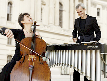 Links im Bild ist Anna Carewe zu sehen wie sie ihr Violoncello spielt. Oli Bott sieht man rechts im Bild stehen, wie er sein Vibraphon spielt. Im Hintergrund ist eine Weiße Hausfassade.