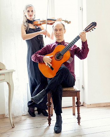 Bild von "duo finesco". Christof Schill sitzt mit seiner Flamencogitarre mittig im Bild auf einem Stuhl. Hinter ihm steht Juliane Winkler mit ihrer Violine.