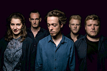 Lukas DeRungs Quintett Band Foto. Die fünf Musiker*innen stehen versetzt und tragen dunkle Kleidung. Sie stehen vor einem schwarzen Hintergrund.