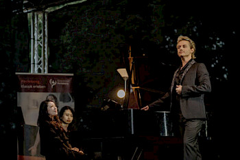 Ein Sänger und 2 Klavierspielerinnen, der Sänger schaut in Fern an.