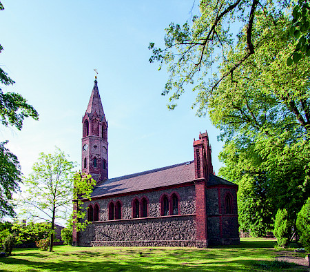 Bild zeigt eine Kirche schräg von vorne mit Bäumen