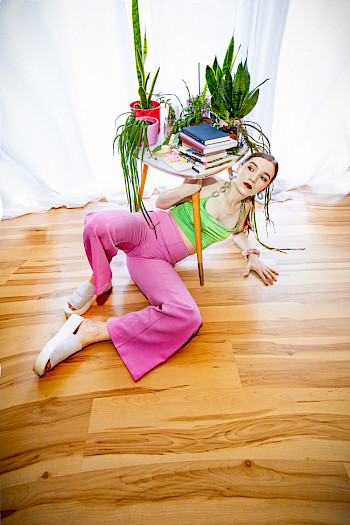 Eine junge Frau mit bunter Kleidung (Rosa <hose, grüne Shirt). Sie liegt unter einem Tisch und hebt ihn hoch.