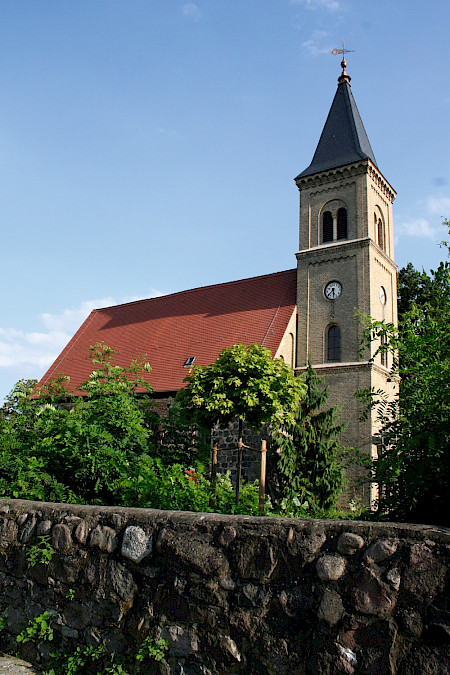 Bild zeigt hinter einer kleinen Mauer eine Kirche mit Kirchenturm und Kirchenuhr