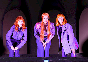 3 Frauen mit orangenen Haaren und lila Kostümen auf einer Bühne