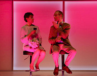 Auf den Hocker sitzen eine Frau und ein Mann in rosaen Licht. Sie haben Wein in den Händen