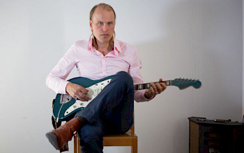 Ein Mann mit blauen Hosen und rosem T- Shirt spielt Guitarre