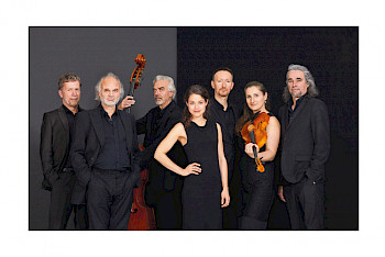 7 MusikerInnen stehen nebeneinander. Sie tragen alle schwarze Klamotte