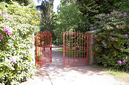 Ein rot gestrichenes Torgitter zu einem Park, eingerahmt von blühenden Rhododendren, ist einladend geöffnet.
