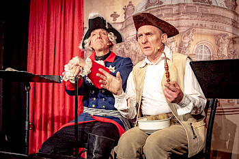 Zwei Schauspieler in historischen Kostümen sitzen nebeneinander auf einer Kutsche: Der preußischer König Friedrich der Zweite neben einem einfachen Bürger, der Kartoffeln auf dem Schoß in einer Schüssel hat.