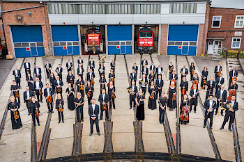 Die Mitglieder eines Orchesters halten ihre Instrumente in den Händen und stehen unter freiem Himmel vor einer Lokhalle