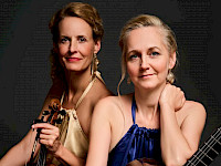 2 Frauen sitzen neben einander. Die eine hält eine Violine.