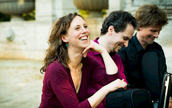 2 Musiker und eine Musikerin stehen auf einer Straße nebeneinander, die Frau lacht