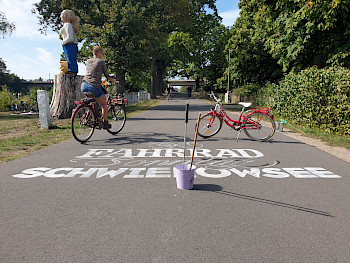 Die Wörter FAHRRAD SCHWIELOWSEE sind mit Kreide auf eine asphaltierte Straße gemalt. Auf der Straße steht ein Eimer, dahinter ein rotes Fahrrad, links auf der Straße eine Frau auf einem Fahrrad, daneben eine Holzstatue