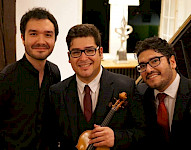 3 junge, südländisch aussehende Männer sehen in die Kamera an und lächeln. Einer hält eine Violine.