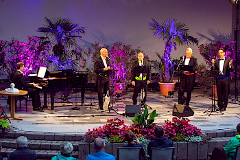 4 Männer im Frack singen auf einer mit Palmen dekorierten Bühne. Neben ihnen ein Mann am Flügel.