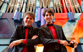 2 junge Männer mit Anzügen und rotem Hemd vor einer farbenprächtigen Orgel