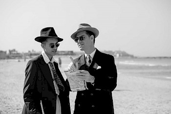 Schwarzweißaufnahme im Stil der 1940er Jahre, die zwei Männer mit Sonnenbrillen, Hüten und Anzug zeigt, die an einem Strand auf einen Stadtplan schauen.