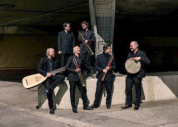 6 schwarz gekleidete Männer mit ihren Blas-, Zupf- und Schlaginstrumenten auf einer Bühne