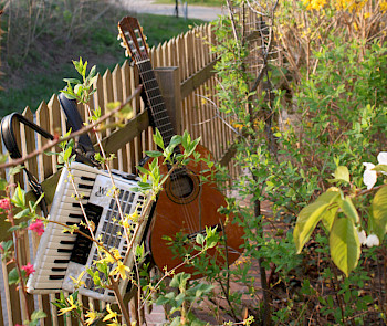 An einem Zaun hängt eine Gitarre und ein Akkordeon, daneben eine Frühlingshecke