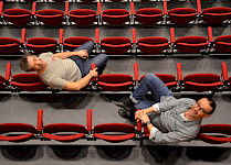 2 Männer sitzen auf Publikum-Stühlen und schauen hoch in eine Kamera über ihnen