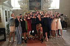 Ein Chor aus jungen, fröhlichen Frauen steht als Gruppe in einer Kirche und zeigt mit den Fingern auf den Fotografen