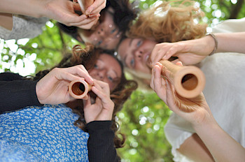 3 Frauen spielen Flöte, die Kamera befindet sich unter den Flöten, so dass der Betrachter in die untere Öffnung der Flöten schaut