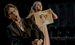 Ein Mann singt mit weit geöffnetem Mund. Im Hintergrund hält eine junge Frau das Bildnis einer Frau aus dem 19. Jahrhundert
