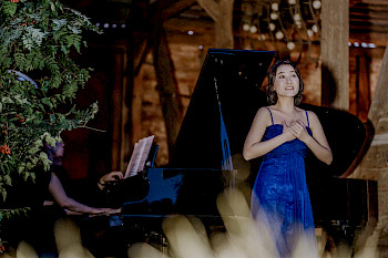 Eine asiatische Sängerin in blauem Abendkleid sing auf einer Bühne, im Hintergrund spielt ein Mann am Flügel