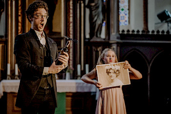 Ein Mann singt mit weit geöffnetem Mund. Im Hintergrund hält eine junge Frau das Bildnis einer Frau aus dem 19. Jahrhundert
