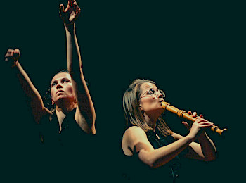 Eine Frau hebt ihre Arme hoch, daneben spielt eine Frau Flöte